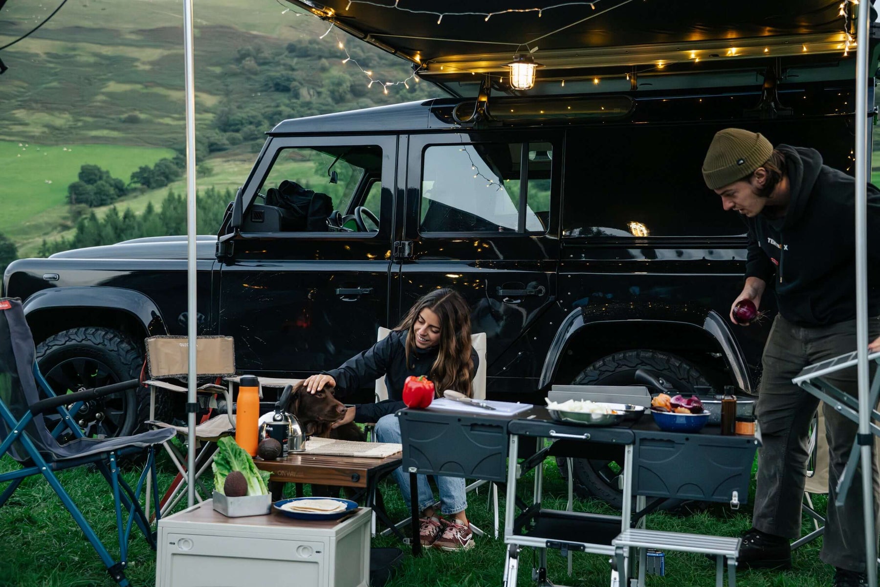 Campo serale con Land Rover Defender, tendalino con luci, cucina da campo e frigo