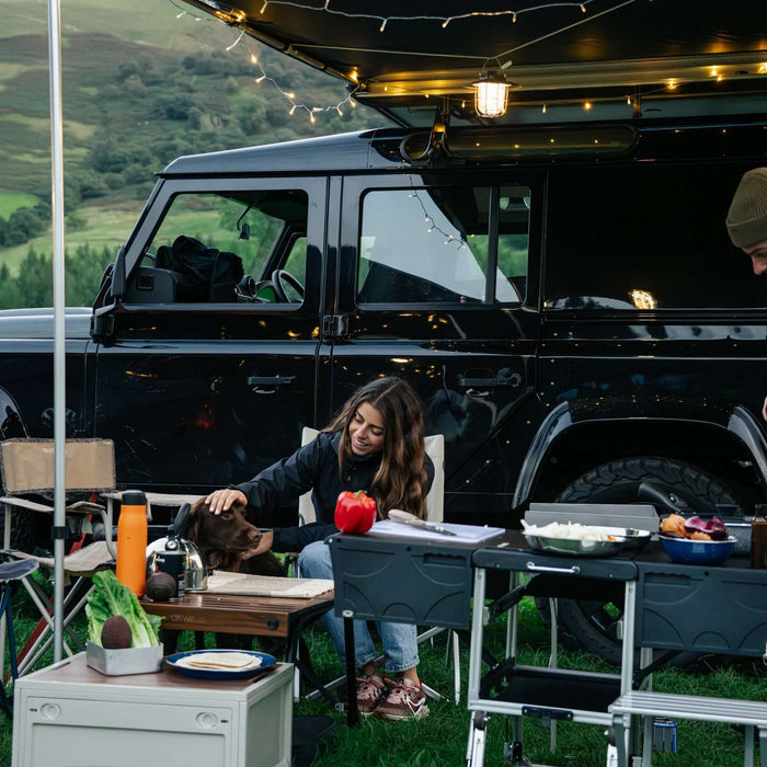 Campo serale con Land Rover Defender, tendalino con luci, cucina da campo e frigo